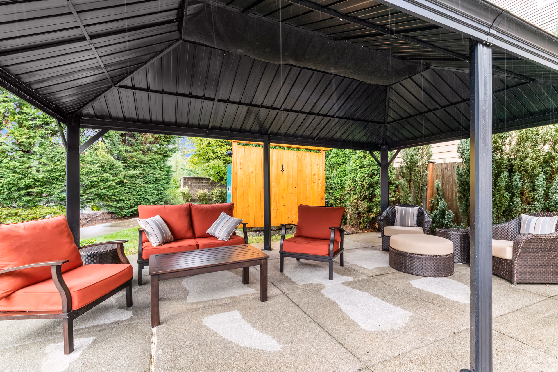 Outdoor covered seating area with red cushioned chairs and a sofa, a dark wooden coffee table, and additional wicker chairs with cushions. The area is surrounded by greenery and a wooden fence, with a concrete floor under the black metal roof structure.