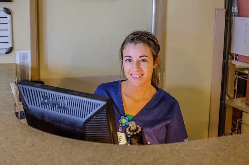 A smiling female healthcare worker wearing a purple scrub top sits behind a reception desk with a computer monitor in front of her inside a nursing and rehabilitation center.