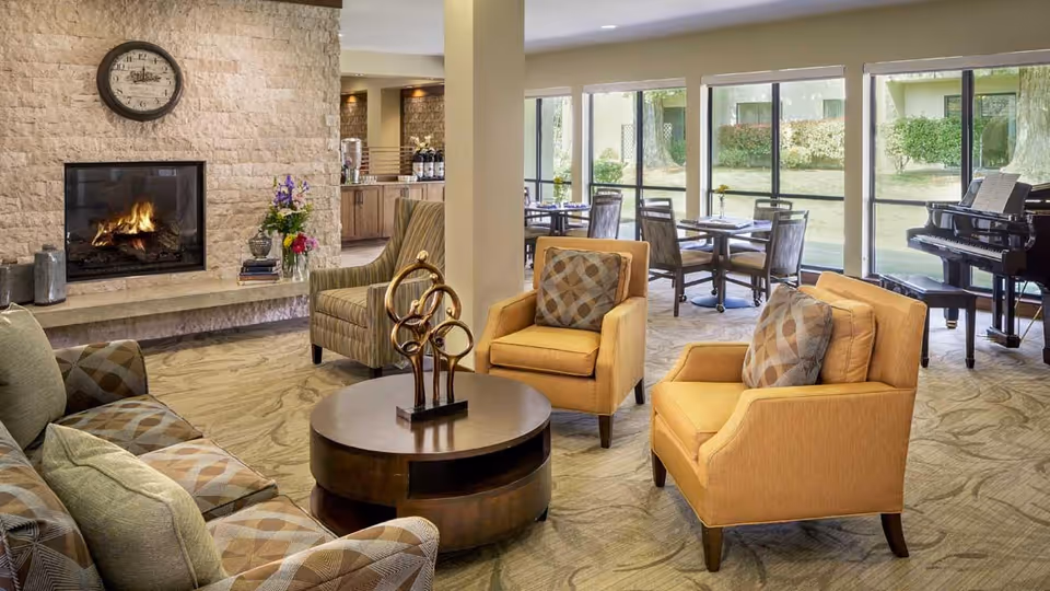 A cozy living room area with a lit fireplace set in a stone wall, a round wooden coffee table with a decorative sculpture, two mustard yellow armchairs with patterned cushions, and a patterned sofa. In the background, there are large windows letting in natural light, several dining tables with chairs, and a black grand piano near the windows.