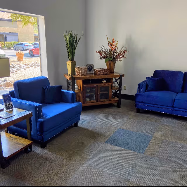 A cozy seating area with two blue upholstered chairs and a matching blue loveseat arranged around a wooden side table and a wooden console table with decorative plants and a clock. A large window shows a parking lot outside.