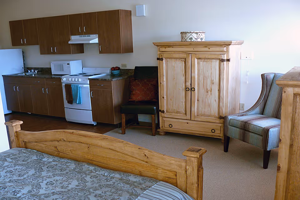 Interior view of a senior living facility room featuring a wooden bed frame with patterned bedding in the foreground, a kitchenette with wooden cabinets, a stove, microwave, and refrigerator along the back wall, a wooden armoire, a striped upholstered chair, and a dark chair with a decorative pillow.