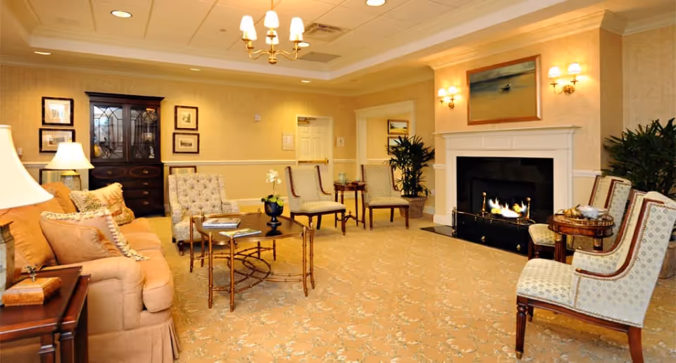 A cozy living room area with a beige sofa, several upholstered chairs, a glass coffee table, and a lit fireplace. The room has warm yellow walls, framed artwork, a chandelier, wall sconces, and potted plants, creating a comfortable and inviting atmosphere.