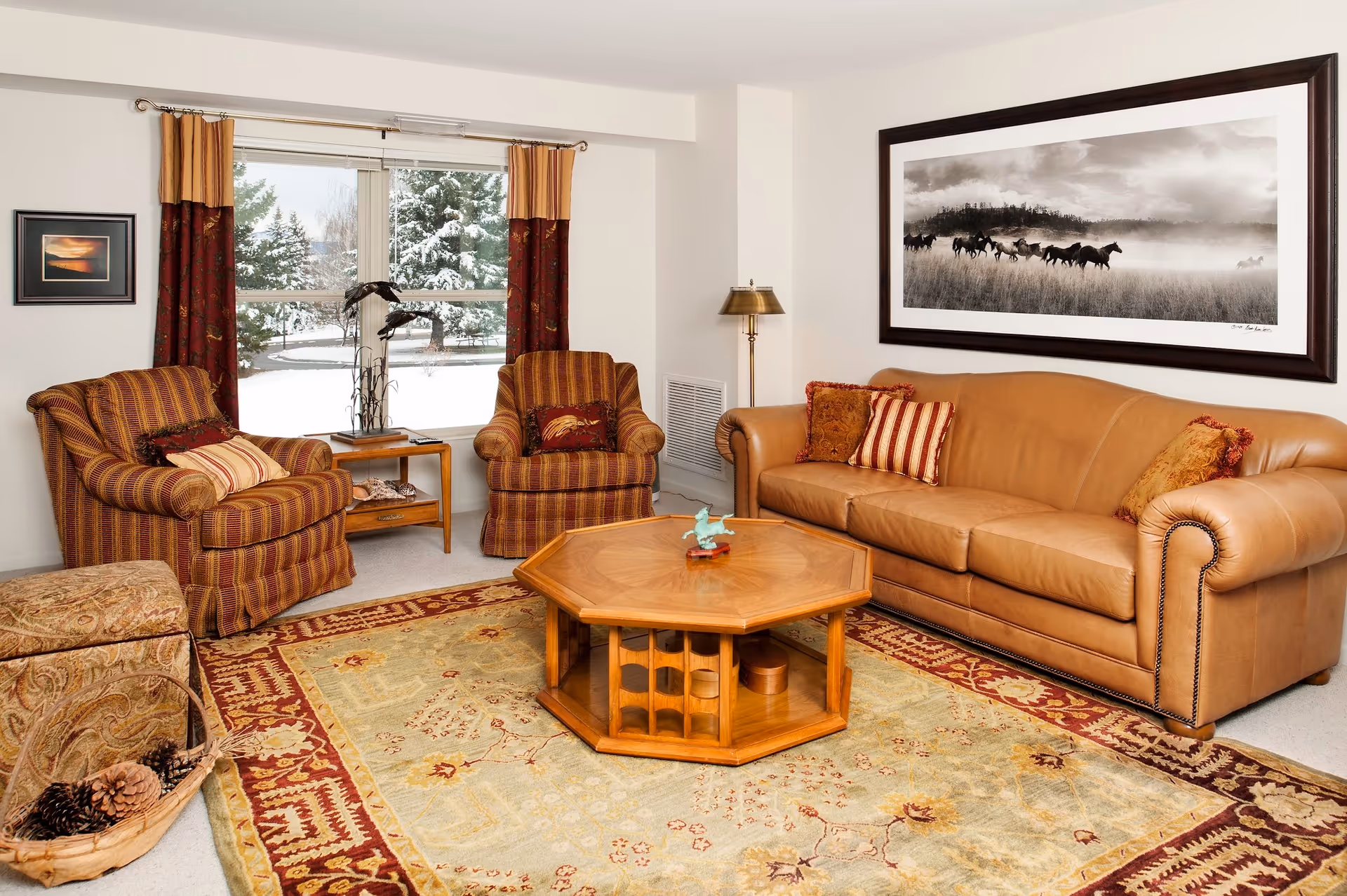 A cozy living room with a tan leather sofa adorned with decorative pillows, two striped armchairs, a wooden octagonal coffee table, and a patterned area rug. A large window with red and gold curtains shows a snowy outdoor scene. A framed black and white photograph of running horses hangs on the wall above the sofa, and a floor lamp stands in the corner.