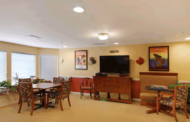 A cozy common area in a senior living facility featuring a wooden table with four floral upholstered chairs, a cushioned bench with a small table, a wooden cabinet with a flat-screen TV mounted above it, and two framed movie posters on the wall. The room has beige walls with a red accent lower half, carpeted floor, and windows with blinds letting in natural light. Several potted plants are placed on the windowsill.