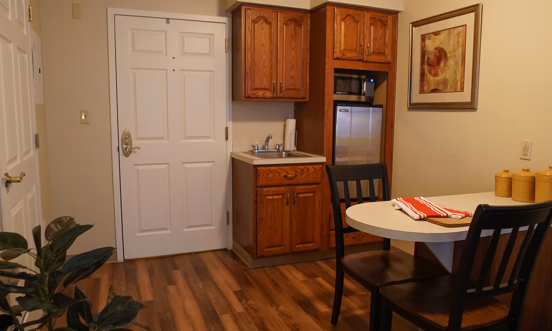 Small kitchen area with wooden cabinets, a sink, a microwave, and a mini refrigerator. A round dining table with two black chairs is set with a folded red and white striped cloth. There is a white door and a framed artwork on the beige wall. A green plant is partially visible in the foreground.