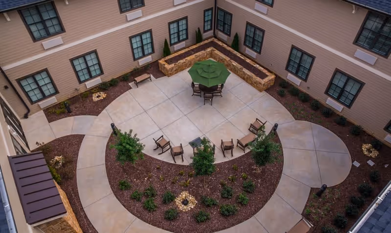 Aerial view of a courtyard in a senior living facility with a circular concrete patio, a table with a green umbrella, several chairs, landscaped garden beds with small shrubs and trees, and surrounding beige building walls with multiple windows.