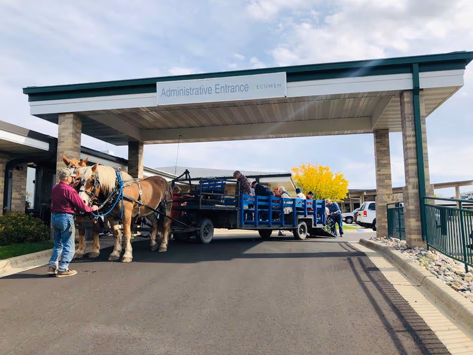 A horse-drawn wagon with several people seated inside is parked under a covered entrance labeled 'Administrative Entrance ECUMEN' at a facility. A man stands beside the horses holding their reins. The scene is outdoors with a clear sky and a tree with yellow leaves in the background.