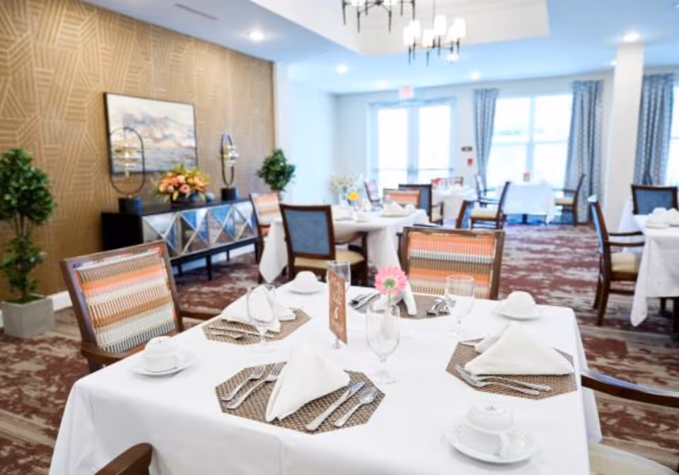 A bright and elegant dining room with tables covered in white tablecloths, set with plates, cups, silverware, and napkins. The room features patterned carpet, decorative chairs with striped upholstery, large windows with curtains, and a sideboard with plants and artwork on the wall.
