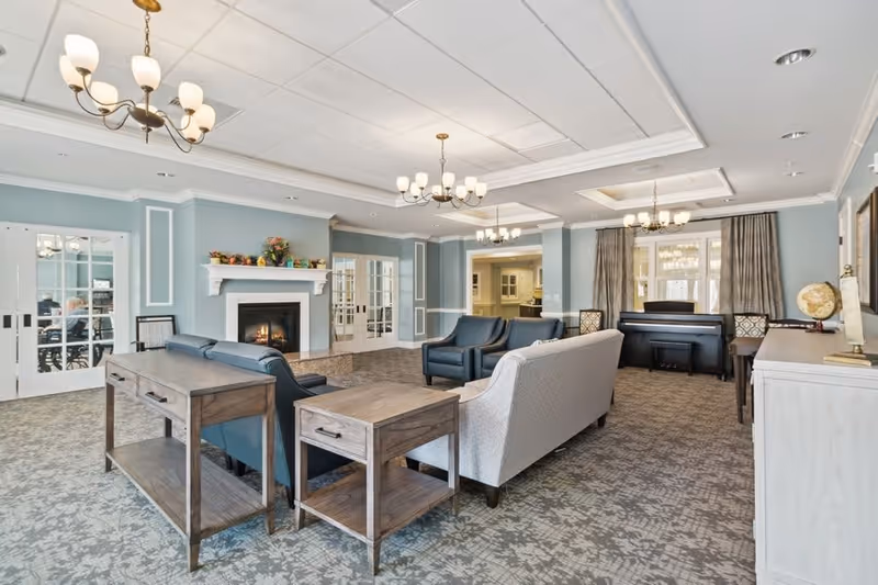 A spacious and well-lit living room with light blue walls, patterned carpet, and multiple chandeliers. The room features a fireplace with a mantel decorated with plants, several sofas and armchairs arranged around a coffee table, a piano near the window with curtains, and wooden side tables. French doors lead to another room in the background.