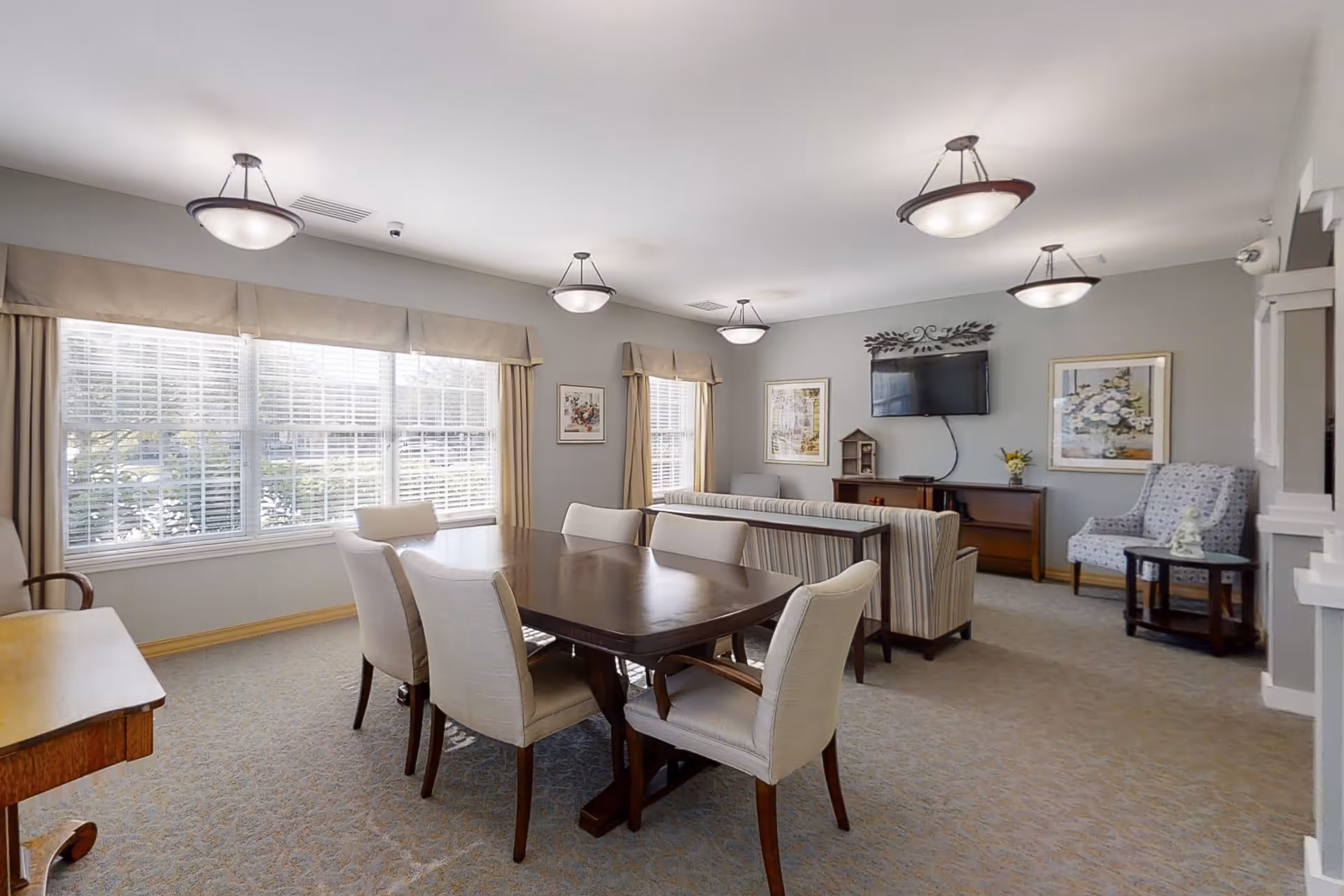A bright and spacious living room area in CountryHouse Residence featuring a wooden dining table with six upholstered chairs, large windows with beige curtains allowing natural light to fill the room, a striped sofa facing a wall-mounted flat-screen TV, framed artwork on the walls, and a patterned armchair next to a small round table.