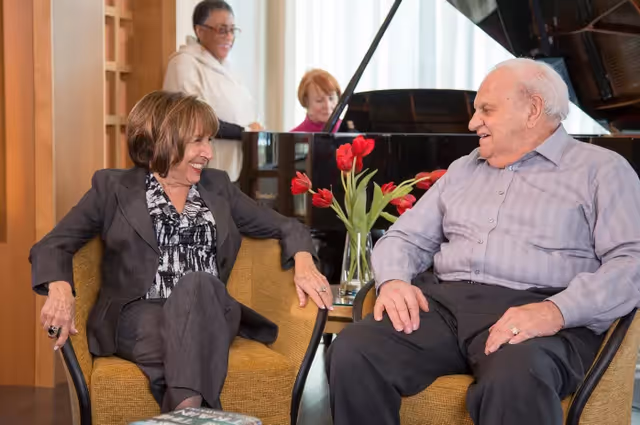 Two elderly people sitting and smiling at each other in a comfortable indoor setting with a vase of red tulips on a table between them. In the background, two women are playing a grand piano.