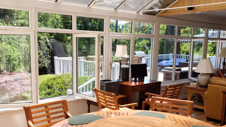 A bright sunroom with large windows and a glass ceiling, featuring wooden chairs around a round wooden table with green placemats. Outside the windows, there is a view of a green garden and a white railing. Inside, there is a wooden desk with a computer monitor and a lamp, and a beige sofa with a side table and lamp.
