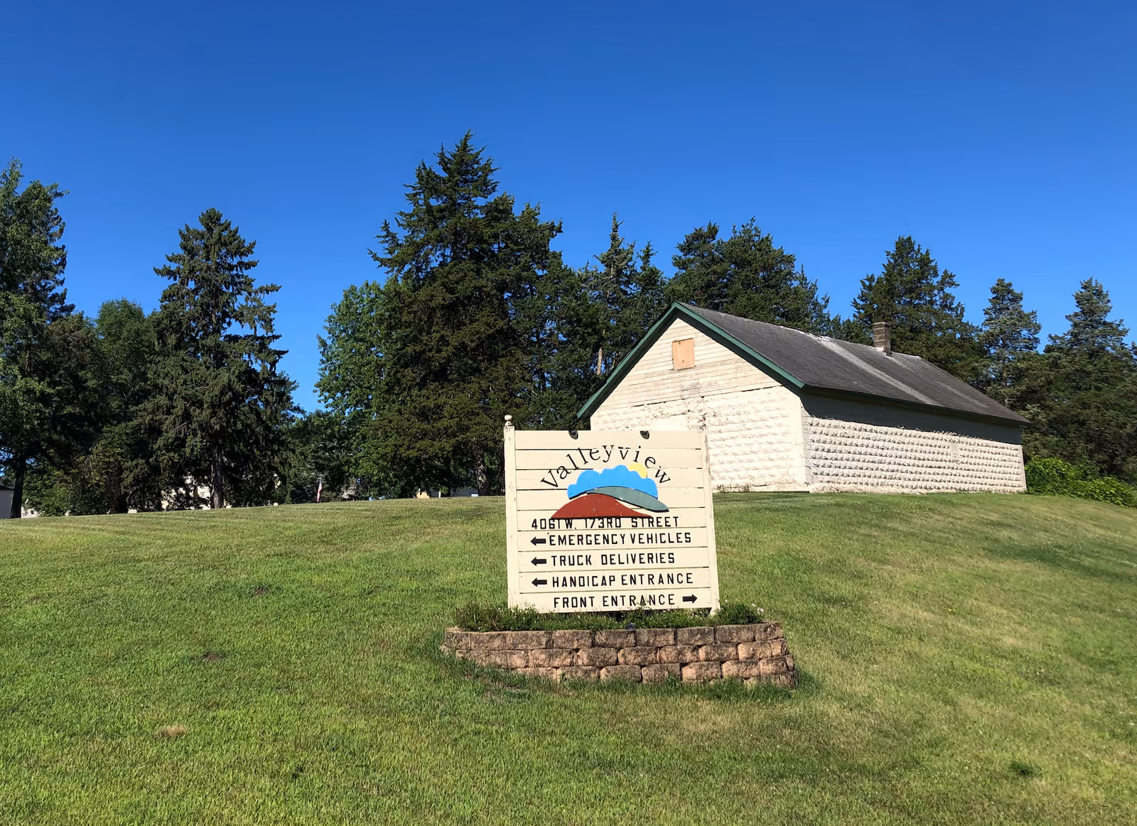 A grassy hill with a sign for Valleyview of Jordan in front of a small white building with a dark roof. The sign includes directions for emergency vehicles, truck deliveries, handicap entrance, and front entrance. Tall trees and a clear blue sky are visible in the background.