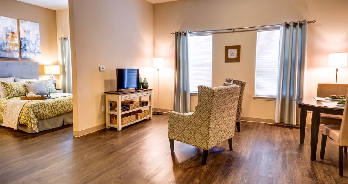 A cozy senior living space with a bedroom on the left featuring a bed with green and beige bedding, a nightstand with a lamp, and artwork above the bed. The main area has two patterned armchairs facing a small TV on a wooden console with baskets and books. There are two windows with light blue curtains and a small dining table with chairs on the right side. The room has warm lighting and wooden flooring.
