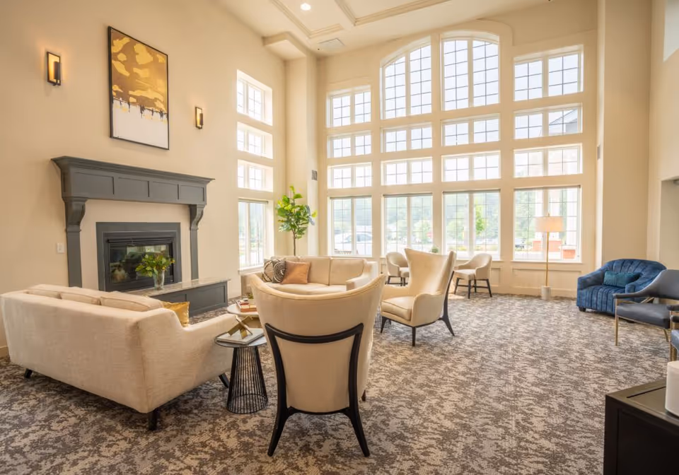Sunlit communal living room with tall arched windows, a seating group arranged around a fireplace, and patterned carpet.
