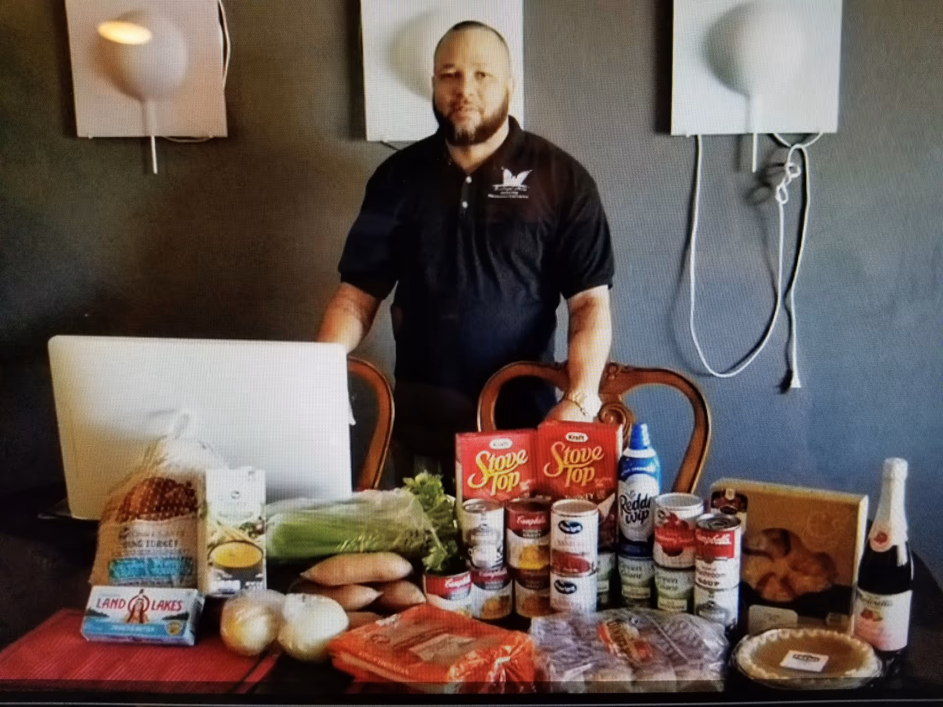 A man stands behind a dining table covered with canned goods, produce and other grocery items in an interior room.