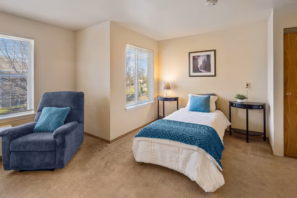 A bright bedroom with beige walls and carpet, featuring a single bed with white bedding and teal accents, a blue armchair with a teal pillow, two small black side tables, a lamp, a framed picture on the wall, and two windows letting in natural light.