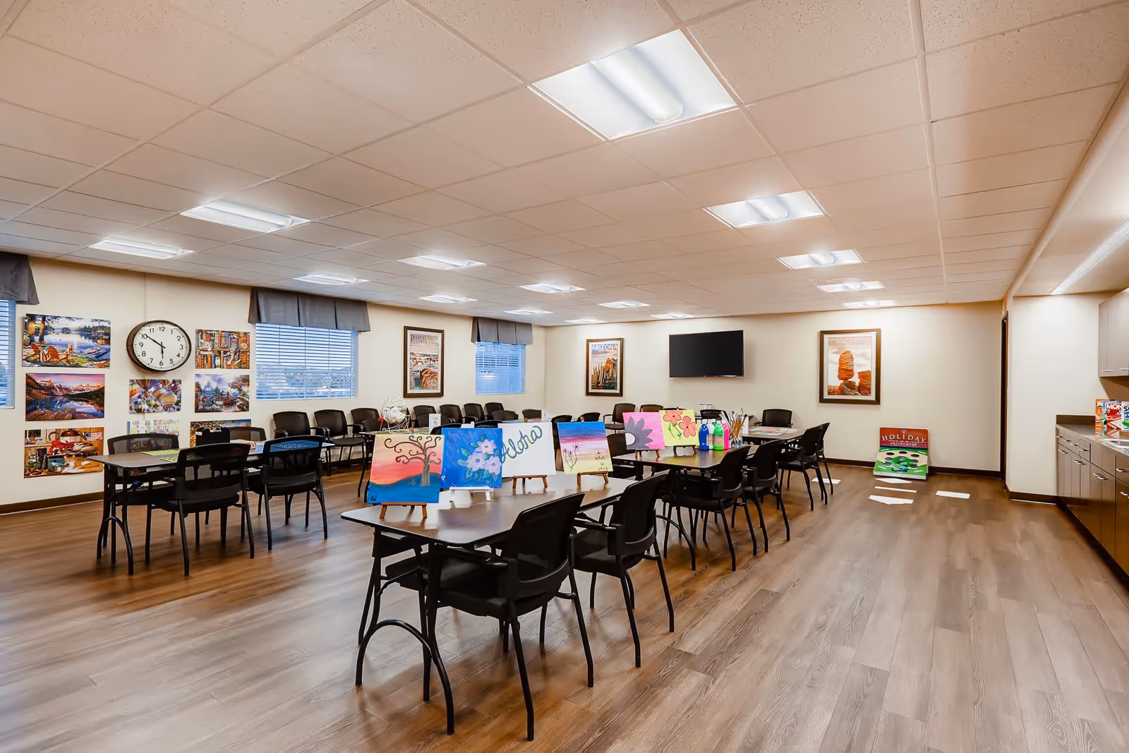 A spacious activity room with several tables and chairs arranged for group activities. On the tables are colorful paintings and art supplies. The room has wood flooring, a wall clock, framed pictures, and a flat-screen TV mounted on the wall. Large windows with valances let in natural light.
