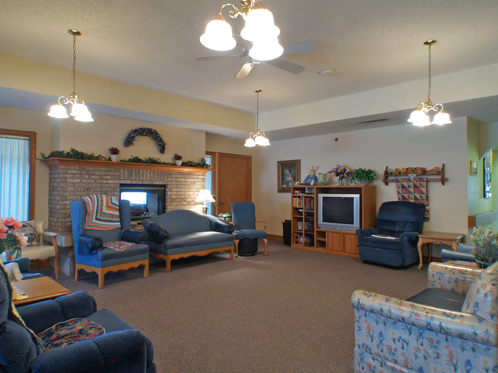 A cozy living room area in a senior living facility with multiple upholstered chairs and a loveseat arranged around a brick fireplace. The room has a ceiling fan with lights, a TV on a wooden stand, bookshelves, and decorative plants and flowers. The walls are light-colored and there are several hanging light fixtures.