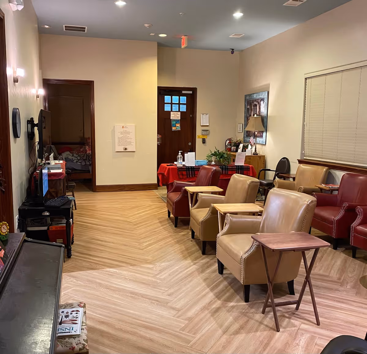 A cozy common living area with several armchairs and small side tables arranged on wood-patterned floors, a TV on the left and a table with plants and supplies by a doorway.