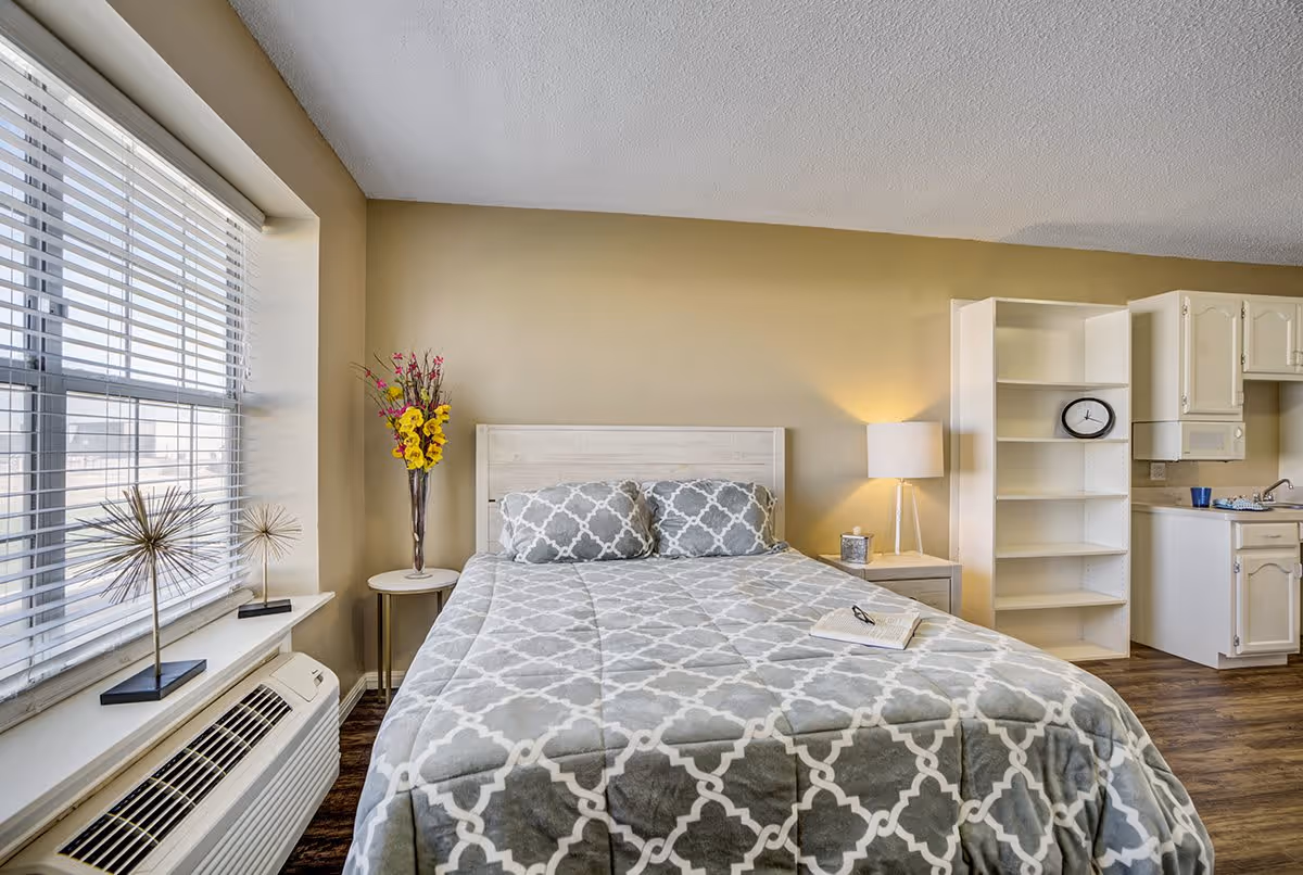 A cozy bedroom with a large window on the left letting in natural light. The bed has a gray and white patterned comforter and two matching pillows. To the left of the bed is a small round table with a tall vase of yellow and pink flowers. To the right is a nightstand with a lamp and a tissue box. Behind the nightstand is a white bookshelf with a clock on one of the shelves. In the background, there is a small kitchenette with white cabinets and a sink. The floor is wooden and the walls are painted beige.