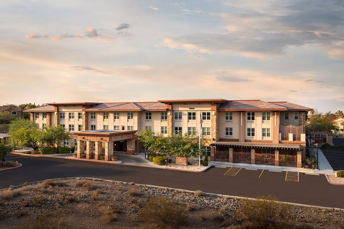 Exterior view of Belmont Village Senior Living Scottsdale, a three-story building with a covered entrance, surrounded by desert landscaping and a parking area under a partly cloudy sky.