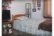 A small bedroom with a single bed covered in light blue patterned bedding and a brown blanket at the foot. Next to the bed is a wooden nightstand with a TV on top and various framed photos and items on the shelves below. On the wall above the bed are several framed pictures and a tall black shelving unit with more photos and decorations. A window with beige curtains is partially visible on the right side.