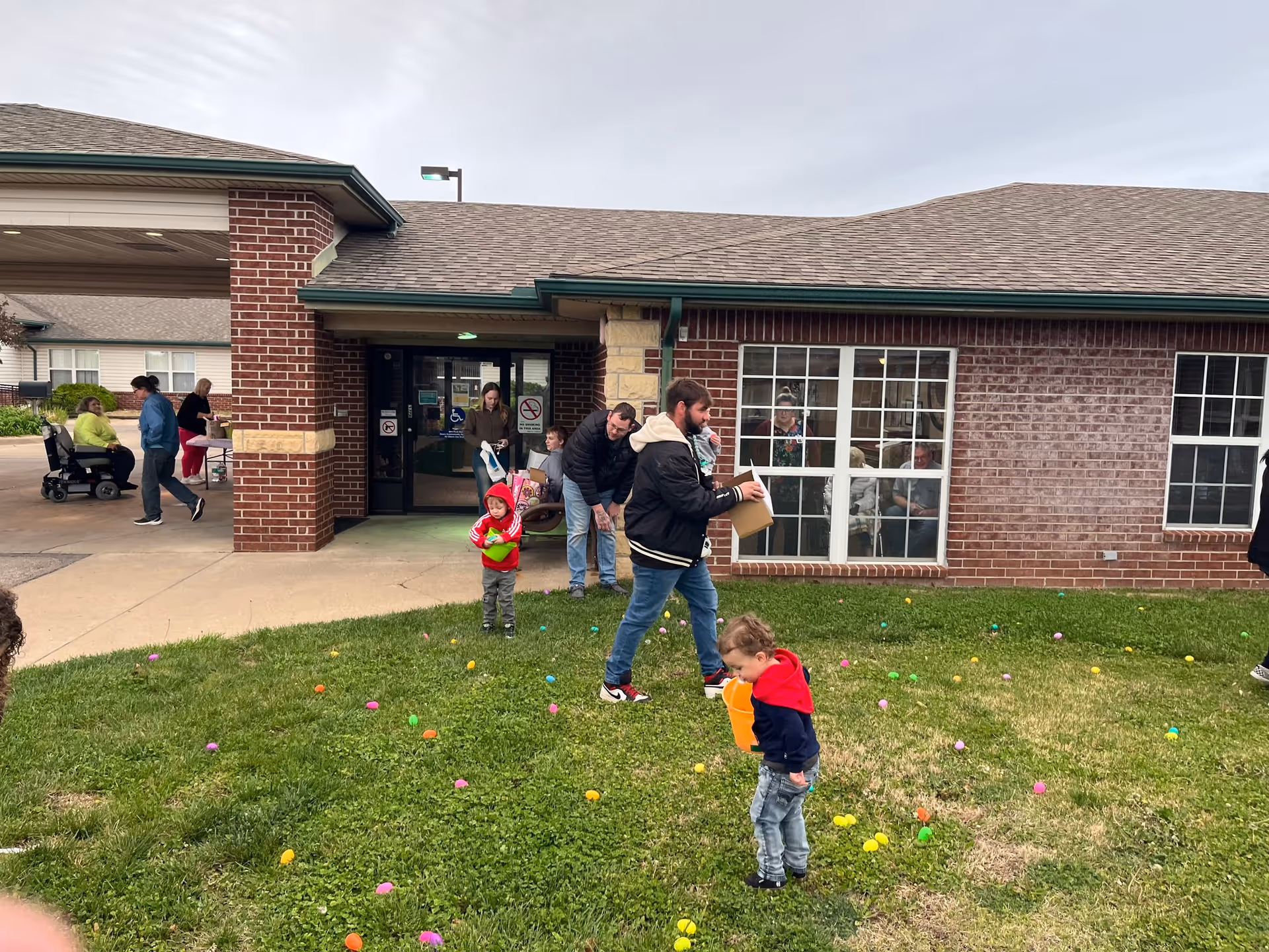 Families and children gather on the grassy front lawn of a brick senior living building, collecting colorful plastic eggs near the entrance.