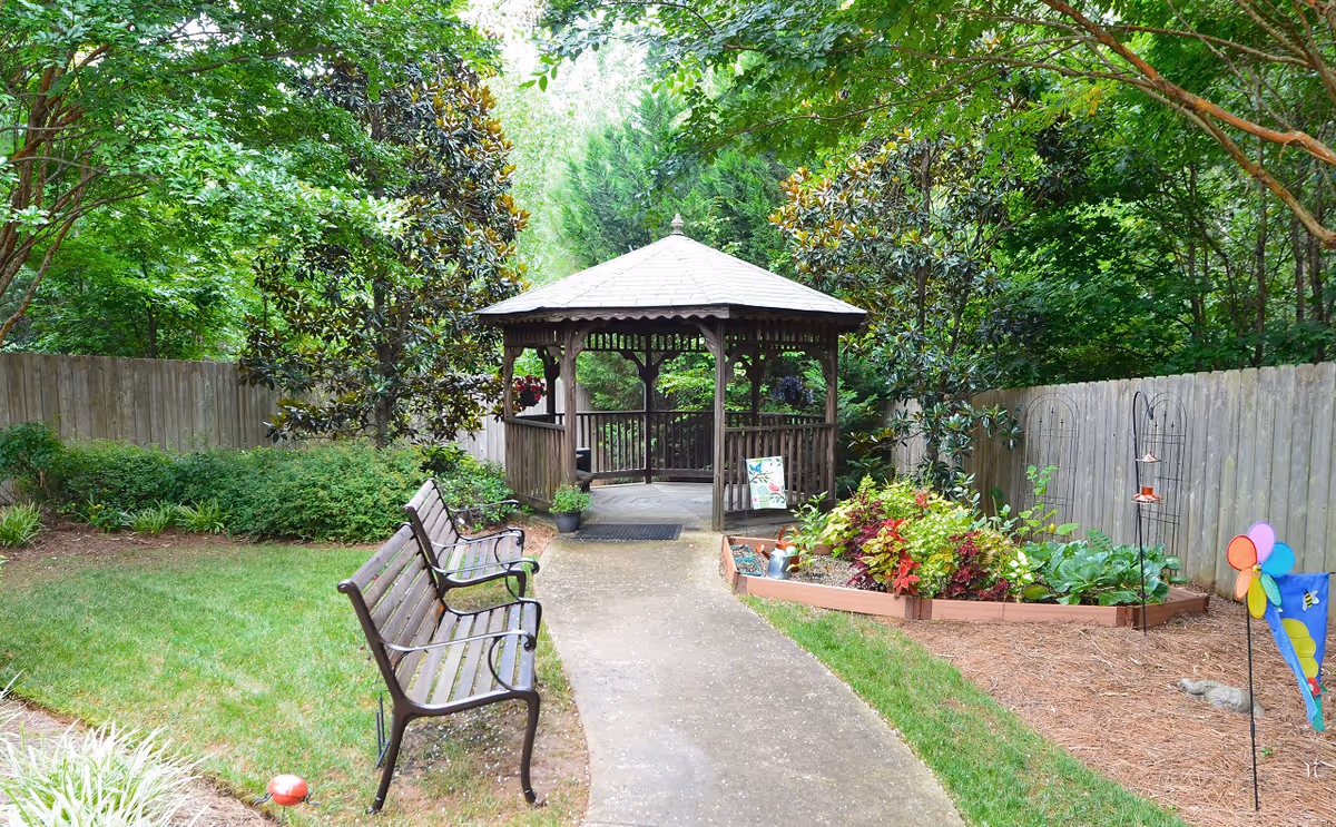 A peaceful outdoor garden area featuring a wooden gazebo at the end of a paved pathway. Two metal and wood benches are placed along the path, surrounded by green grass, bushes, and trees. There is a raised garden bed with various plants and flowers on the right side, along with colorful garden decorations including a pinwheel and a small flag.