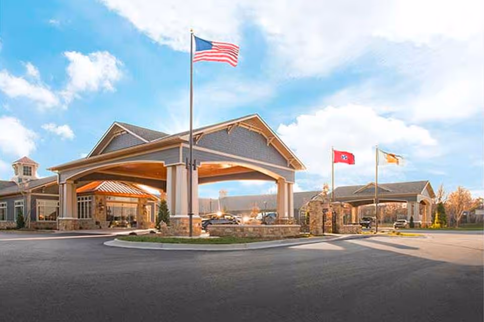 Front exterior view of Life Care Center of East Ridge showing a covered entrance with three flagpoles flying the American flag, a state flag, and another flag, under a partly cloudy sky.