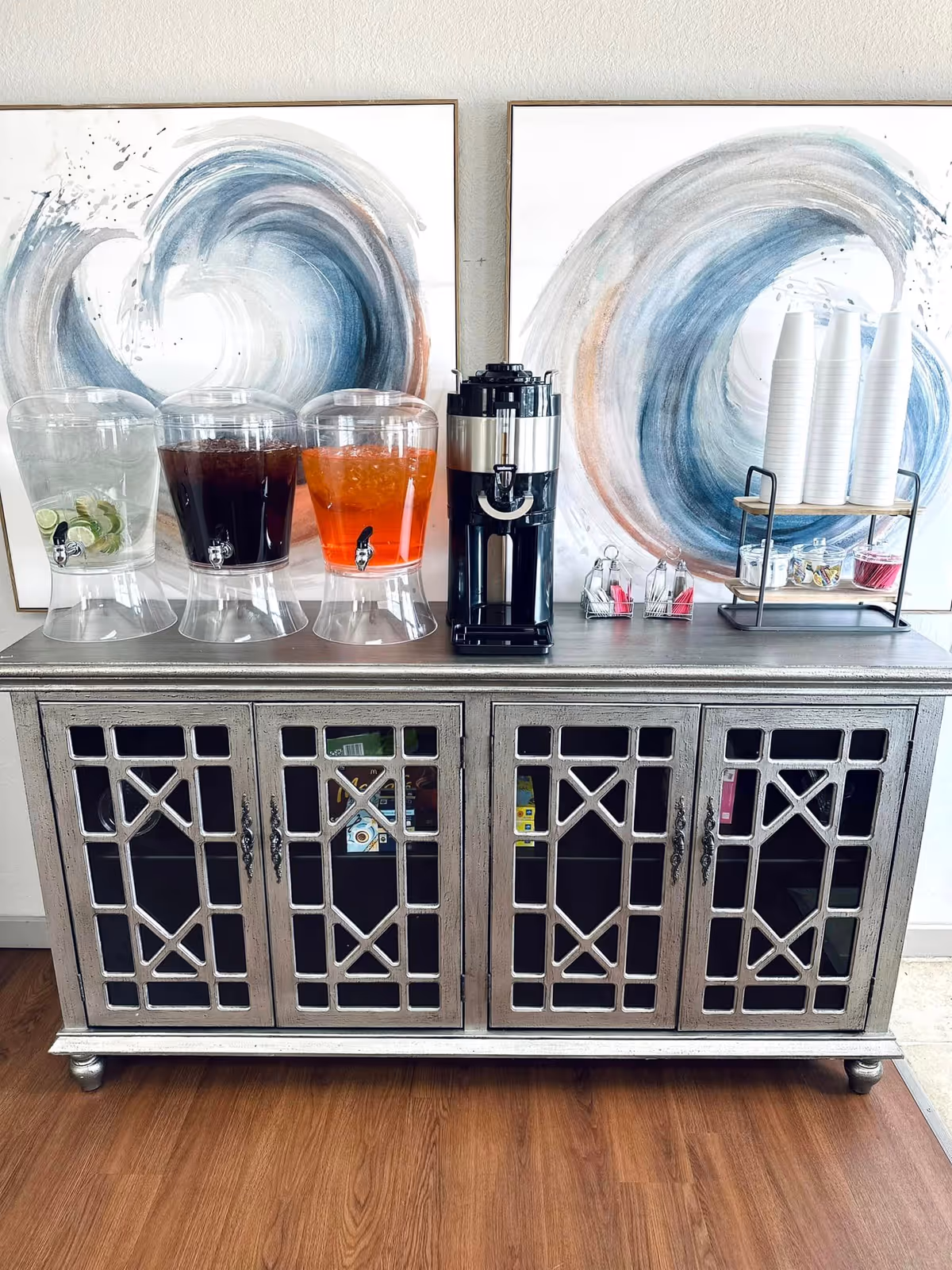 A beverage station with three drink dispensers, a coffee urn, stacked disposable cups and condiments arranged on a decorative cabinet beneath abstract wall art.