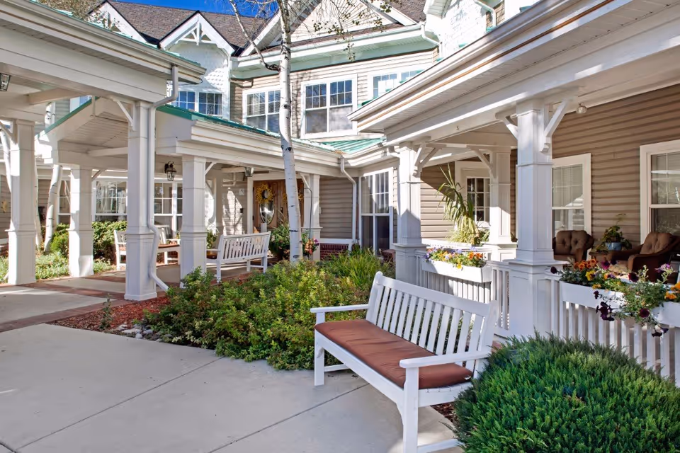 Outdoor seating area at Sunrise at Orchard featuring white wooden benches with brown cushions, surrounded by green shrubs and colorful flowers under a covered walkway attached to a beige building with white trim and multiple windows.