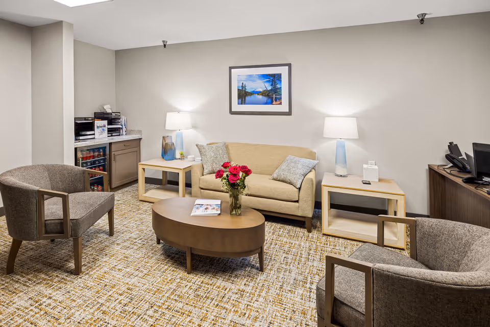A cozy senior living community lounge area with a beige sofa, two gray armchairs, a wooden oval coffee table with a vase of red roses, two side tables each with a lamp, a framed landscape photograph on the wall, and a small counter with a mini fridge and office supplies.