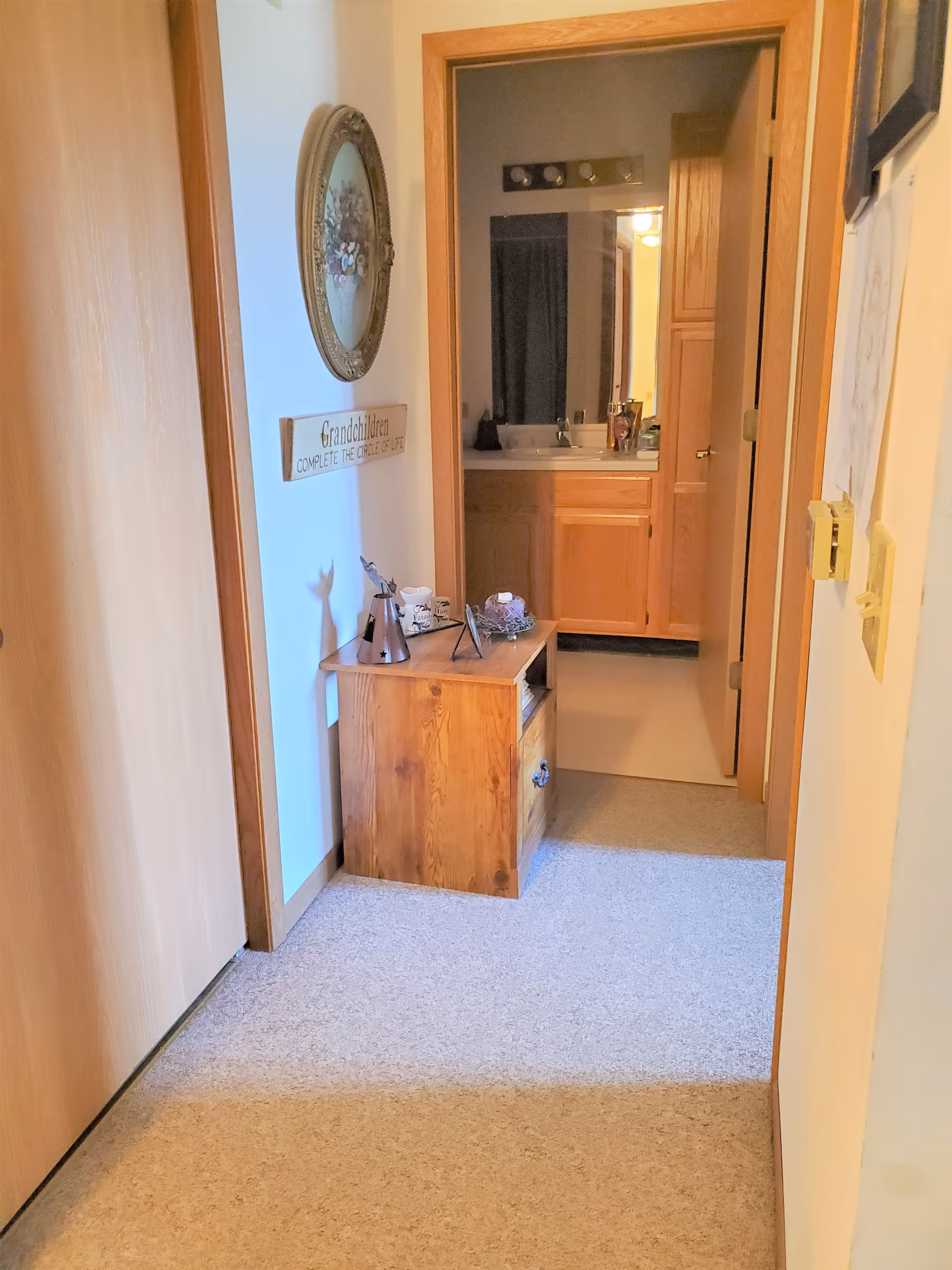 A hallway with light-colored carpet and wooden doors leading to a bathroom with wooden cabinets and a sink. A small wooden cabinet with decorative items sits against the wall, which also has a round floral wall decoration and a sign that reads 'Grandchildren complete the circle of life.'