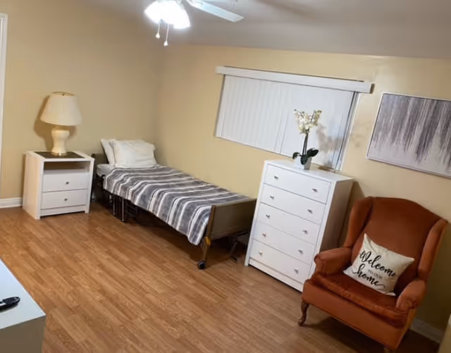 A senior living facility bedroom with a single bed covered in a gray and white striped blanket, a white pillow, and a white nightstand with a lamp. There is a white chest of drawers with a potted orchid on top, a brown upholstered armchair with a 'Welcome Home' pillow, a window with vertical blinds, and a piece of abstract wall art. The floor is wooden and the walls are beige.