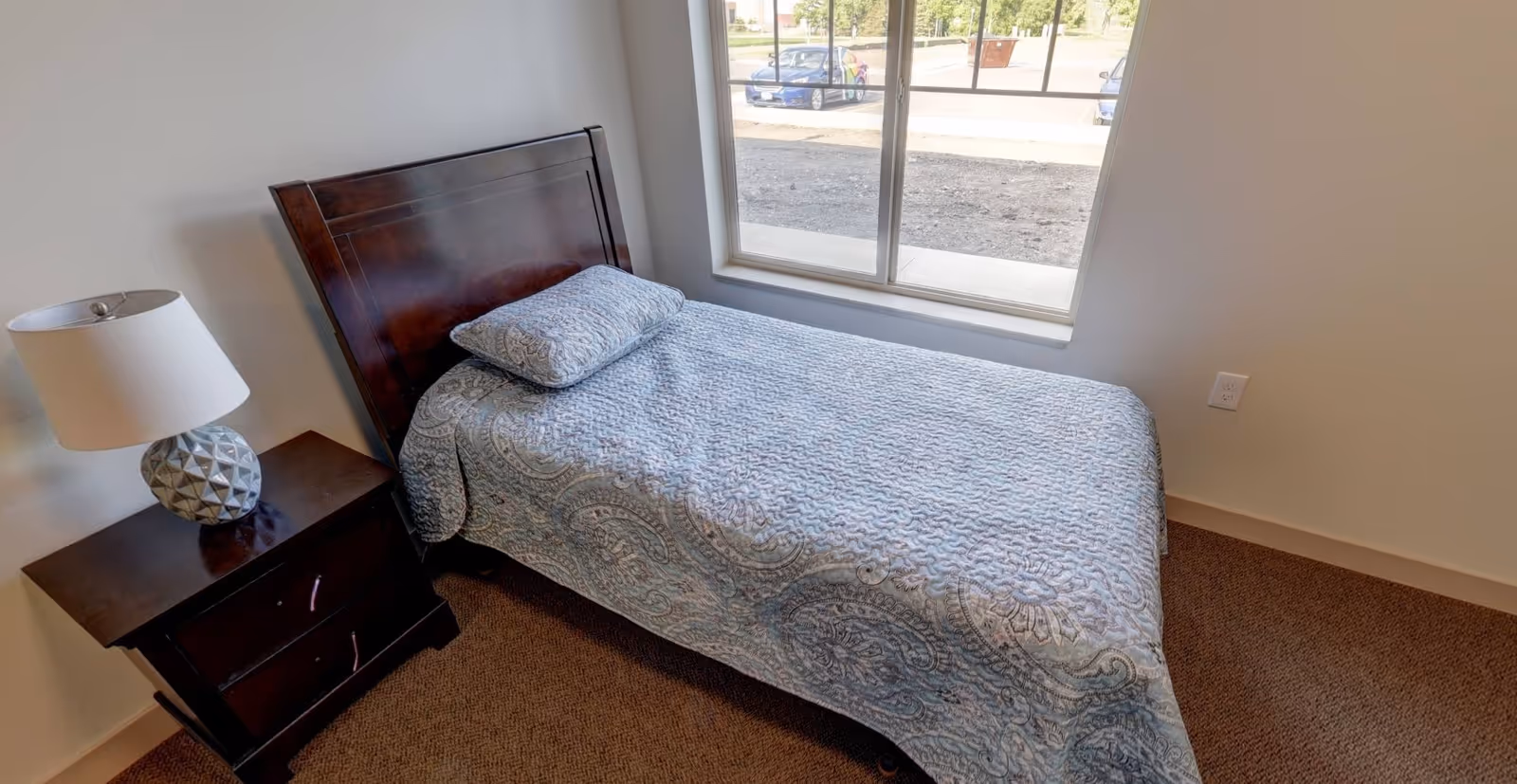 A small bedroom with a single bed covered in a light blue patterned quilt and matching pillow. Next to the bed is a dark wooden nightstand with two drawers and a decorative lamp on top. A large window behind the bed lets in natural light and shows a view of parked cars outside.