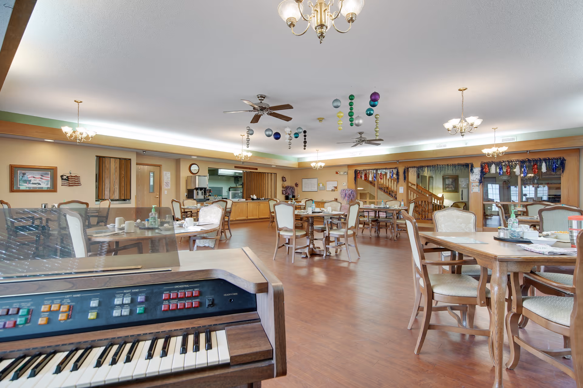 A spacious dining room with several wooden tables and cushioned chairs arranged neatly. The room features a wooden floor, ceiling fans, and multiple chandeliers. There is a keyboard instrument in the foreground, and colorful hanging decorations from the ceiling. The back wall has a counter area and a doorway leading to another room, with festive garlands hanging above a large window or mirror.
