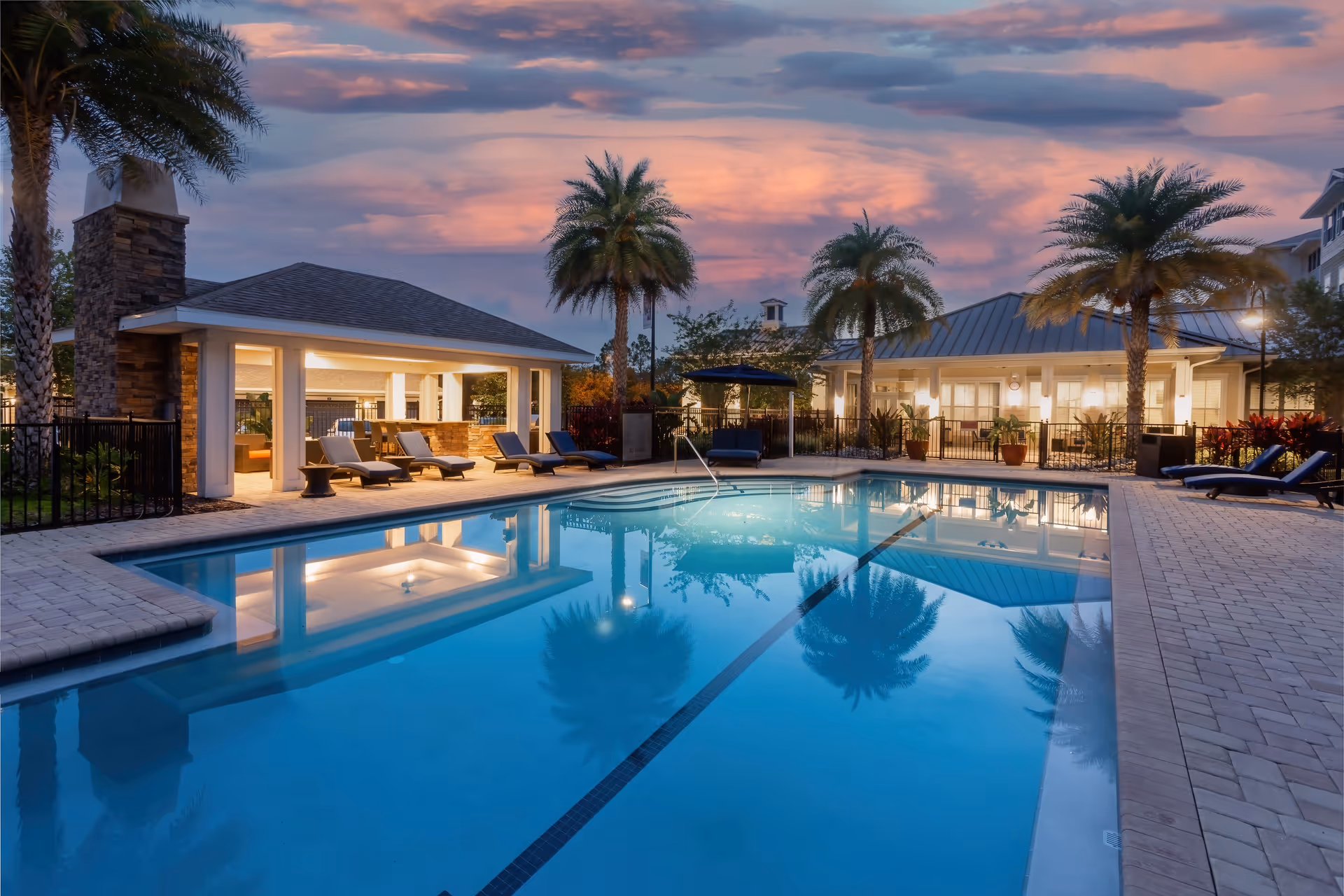 Outdoor swimming pool with lounge chairs, palm trees, and an illuminated clubhouse at sunset.