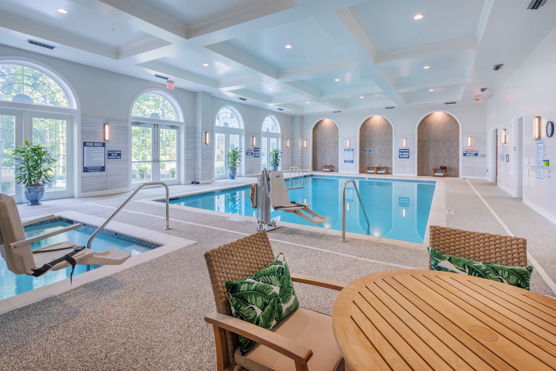 Bright indoor swimming pool and hot tub area with seating, pool lifts, and large arched windows.