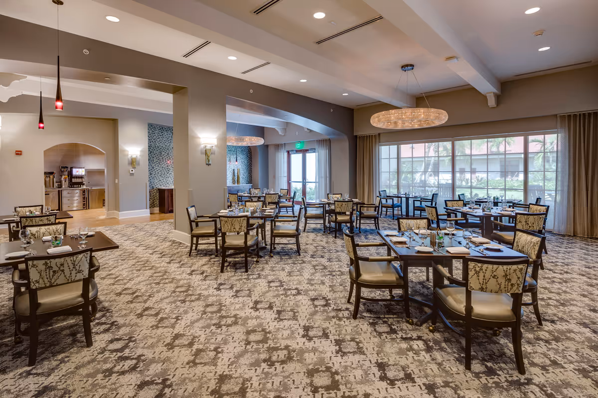 Spacious dining room with multiple tables and chairs arranged neatly. Each table is set with plates, glasses, and napkins. Large windows on one side allow natural light to fill the room, and a modern chandelier hangs from the ceiling. The room has a patterned carpet and neutral-colored walls with soft lighting fixtures.
