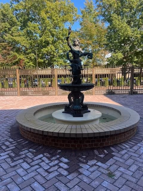 Outdoor circular brick fountain with a statue of a woman holding an object, surrounded by a paved area and metal fencing, with trees and greenery in the background under a clear sky.