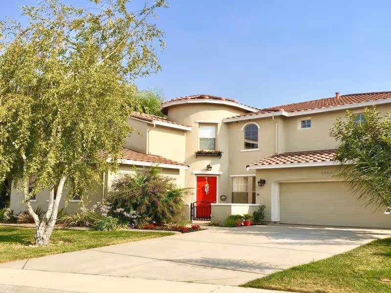 Exterior view of a two-story residential building with beige walls and a red front door. The house has a tiled roof and a two-car garage. There is a driveway leading up to the garage and front door, with green grass and trees on either side. The sky is clear and blue.