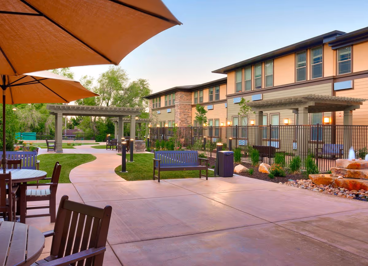 Outdoor courtyard with patio seating and umbrellas, benches and pergolas in front of a two-story senior living building.