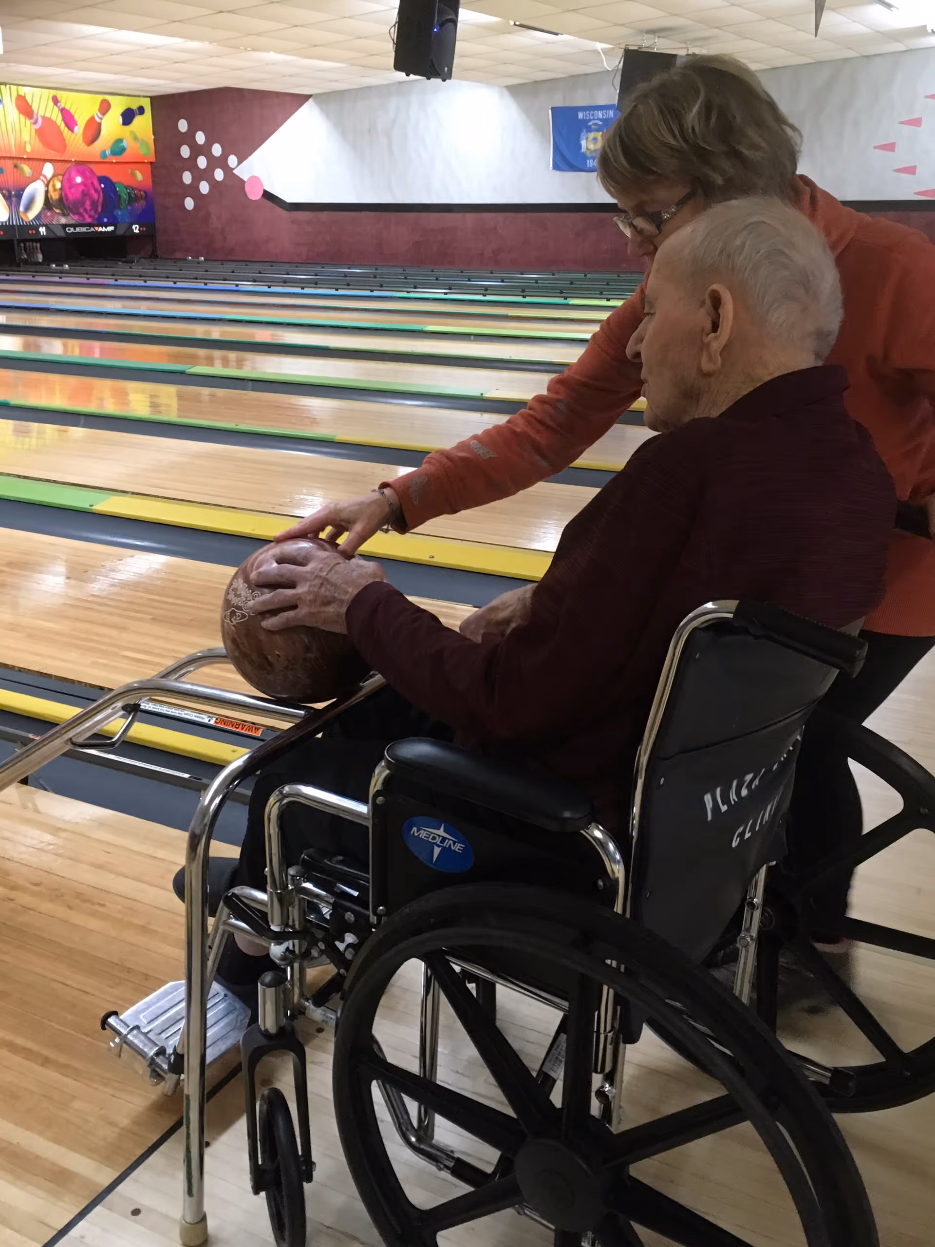 An elderly man in a wheelchair is holding a bowling ball with assistance from a woman standing beside him at a bowling alley. Several bowling lanes are visible in the background.