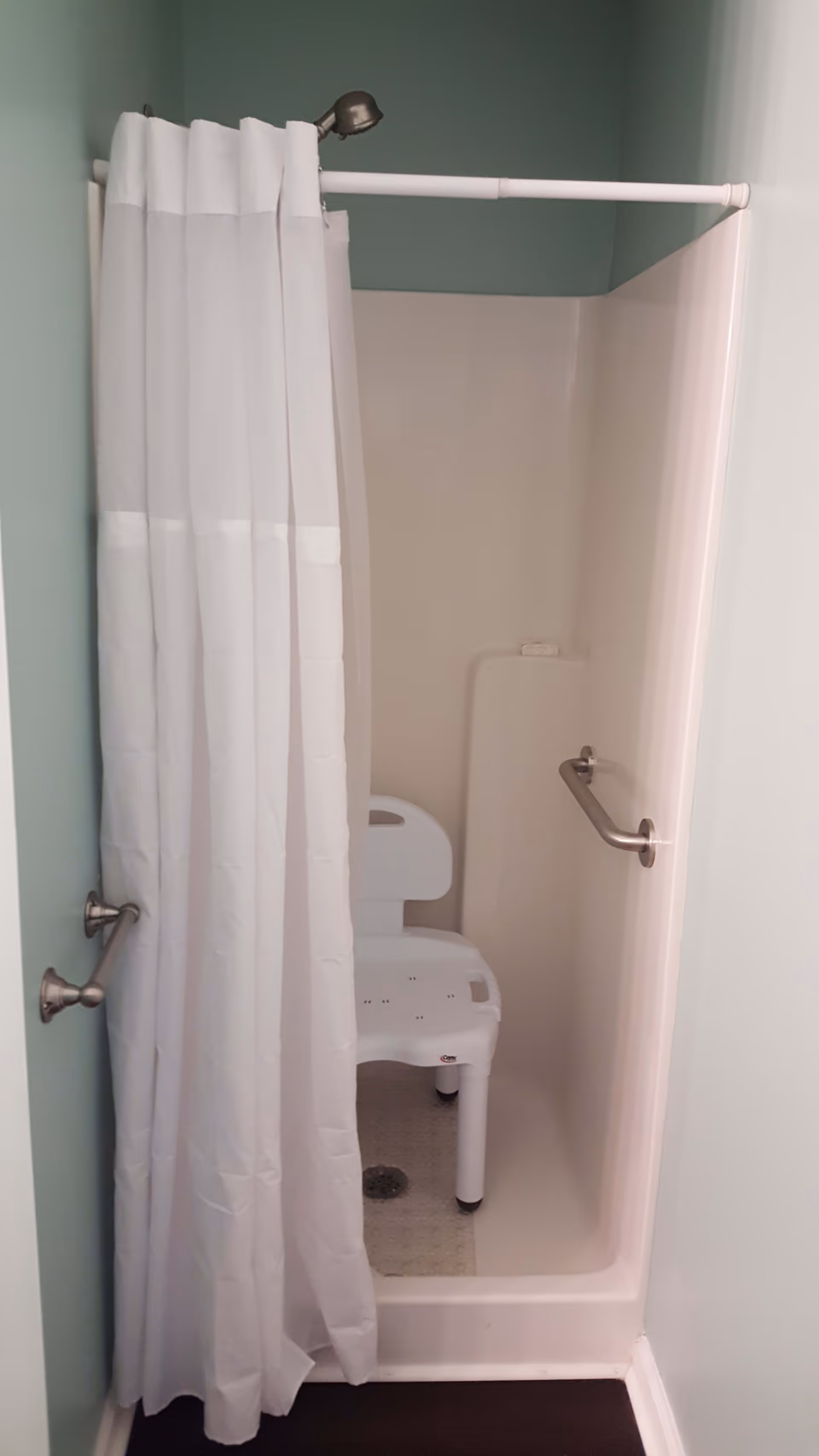 A small shower stall with a white shower curtain partially drawn to the side. Inside the shower is a white plastic shower chair with a backrest. There are two metal grab bars, one on the right wall inside the shower and one on the left wall outside the shower. The walls are painted light green and the floor outside the shower is dark wood.