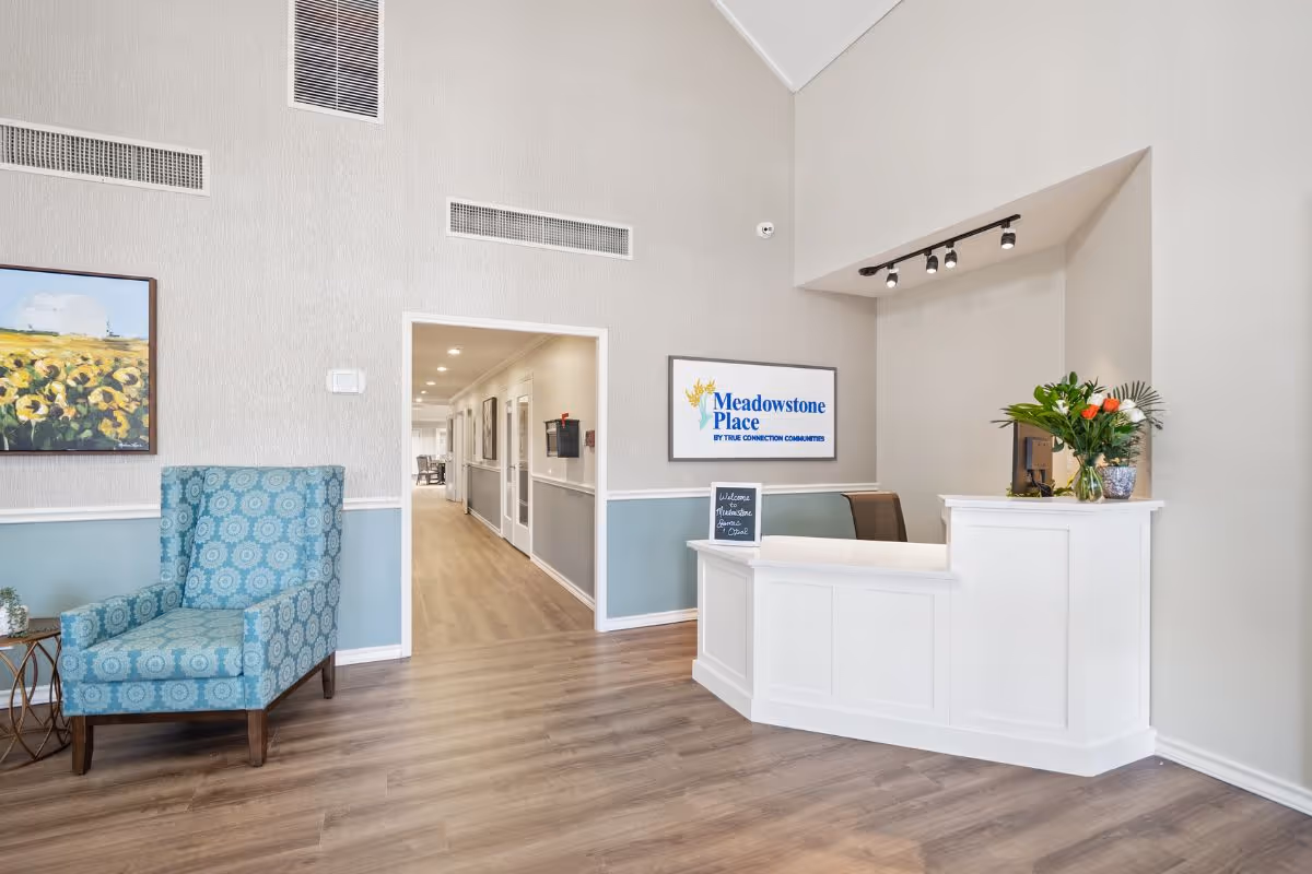 Reception area of Meadowstone Place featuring a white reception desk with a computer and a vase of flowers, a blue patterned armchair, a painting of sunflowers on the wall, and a hallway leading further into the facility.