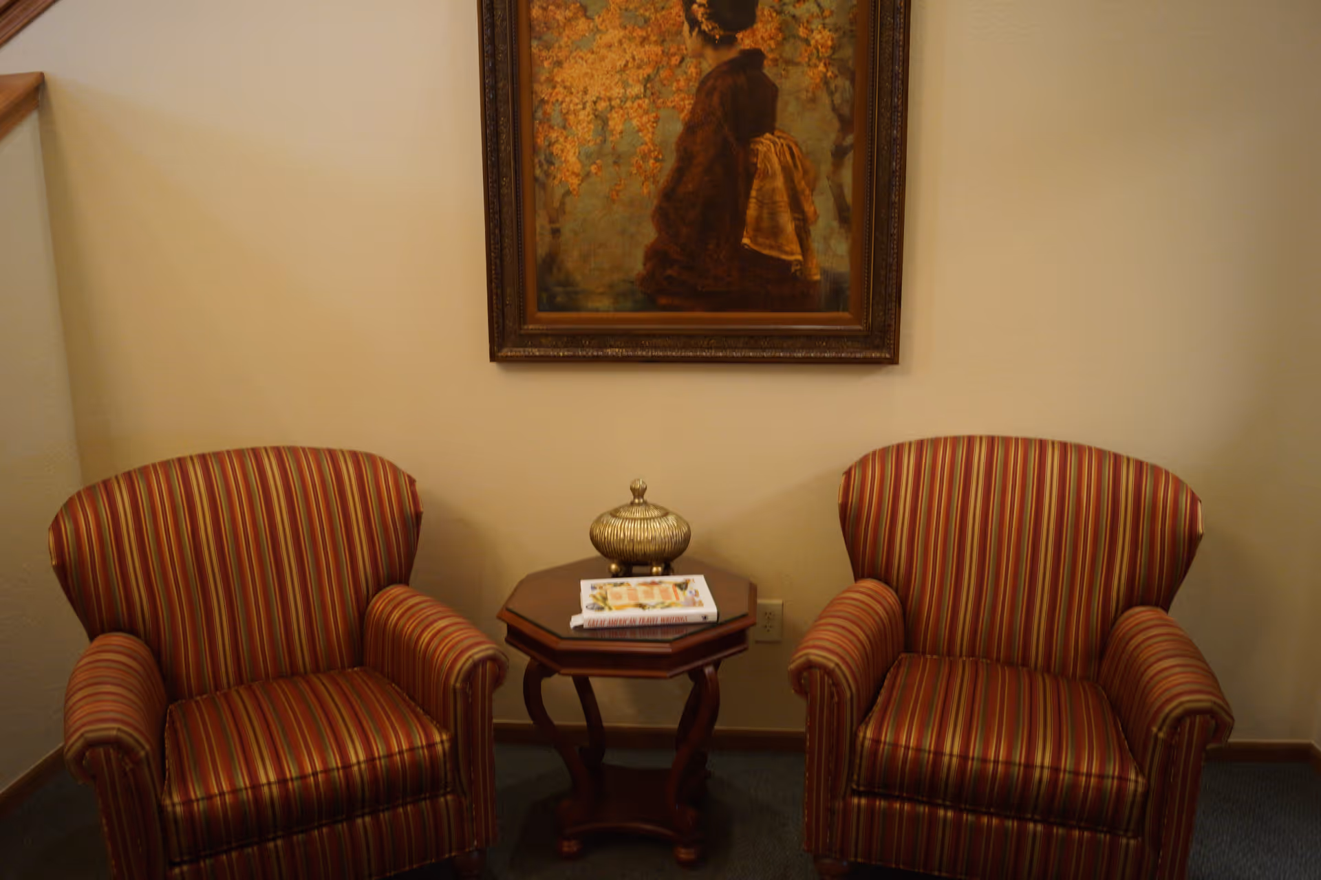 Two striped upholstered armchairs with a small wooden side table between them. On the table is a decorative container and a magazine. Above the chairs hangs a framed painting of a woman in traditional attire with an orange floral background.