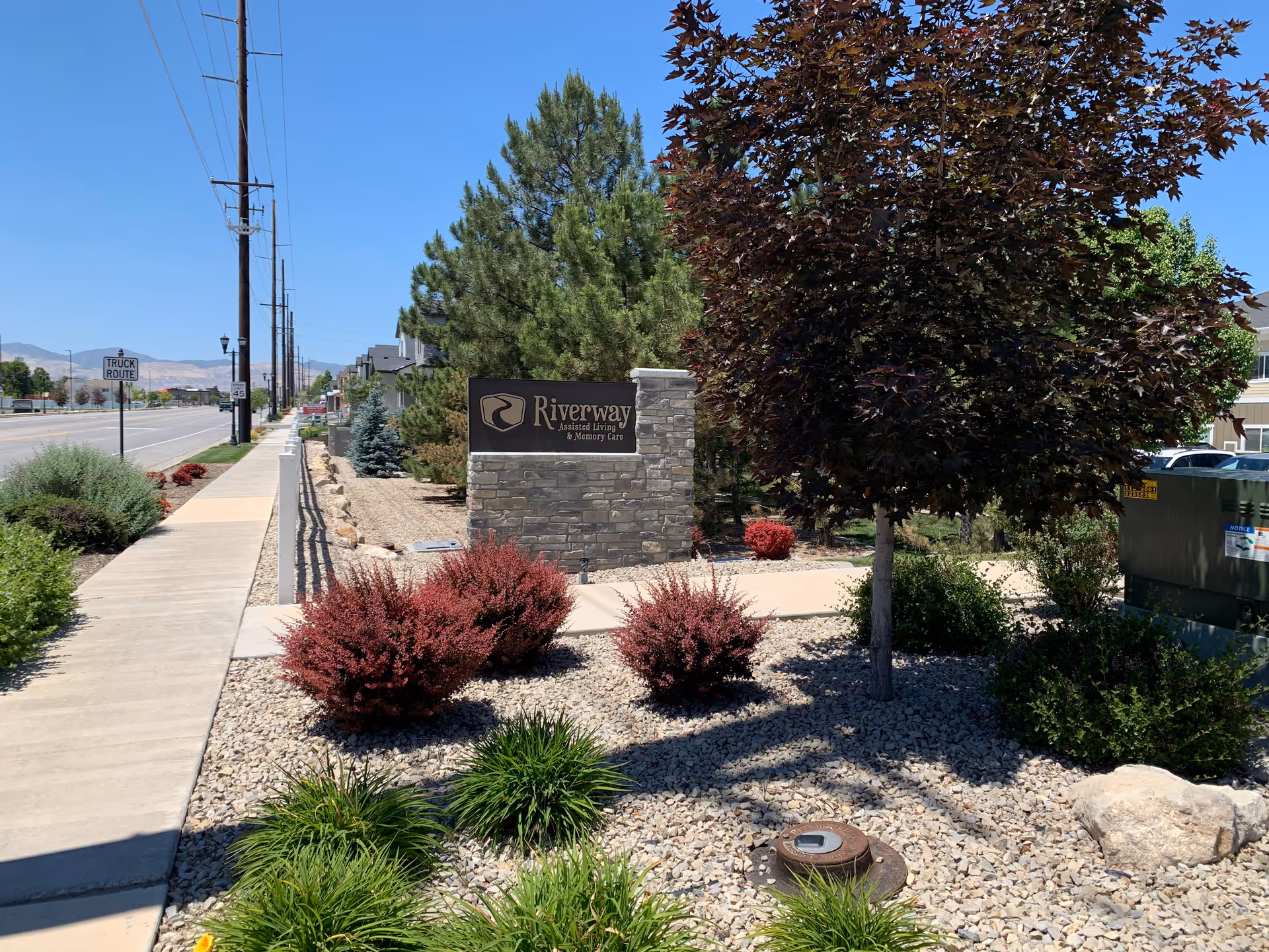 Outdoor view of the entrance area to Riverway Assisted Living and Memory Care facility, featuring a stone sign with the facility's name surrounded by landscaped bushes, trees, and rocks along a sidewalk next to a street with utility poles and traffic signs.