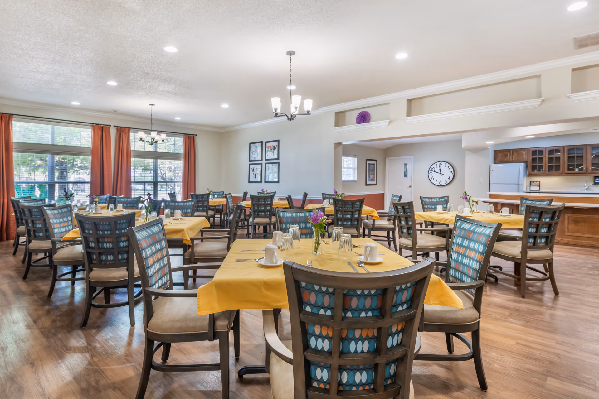 A bright dining room with multiple tables covered in yellow tablecloths, each set with cups, glasses, and small flower vases. The room has wooden chairs with patterned backs, large windows with red curtains, and a kitchen area with wooden cabinets and a refrigerator in the background.