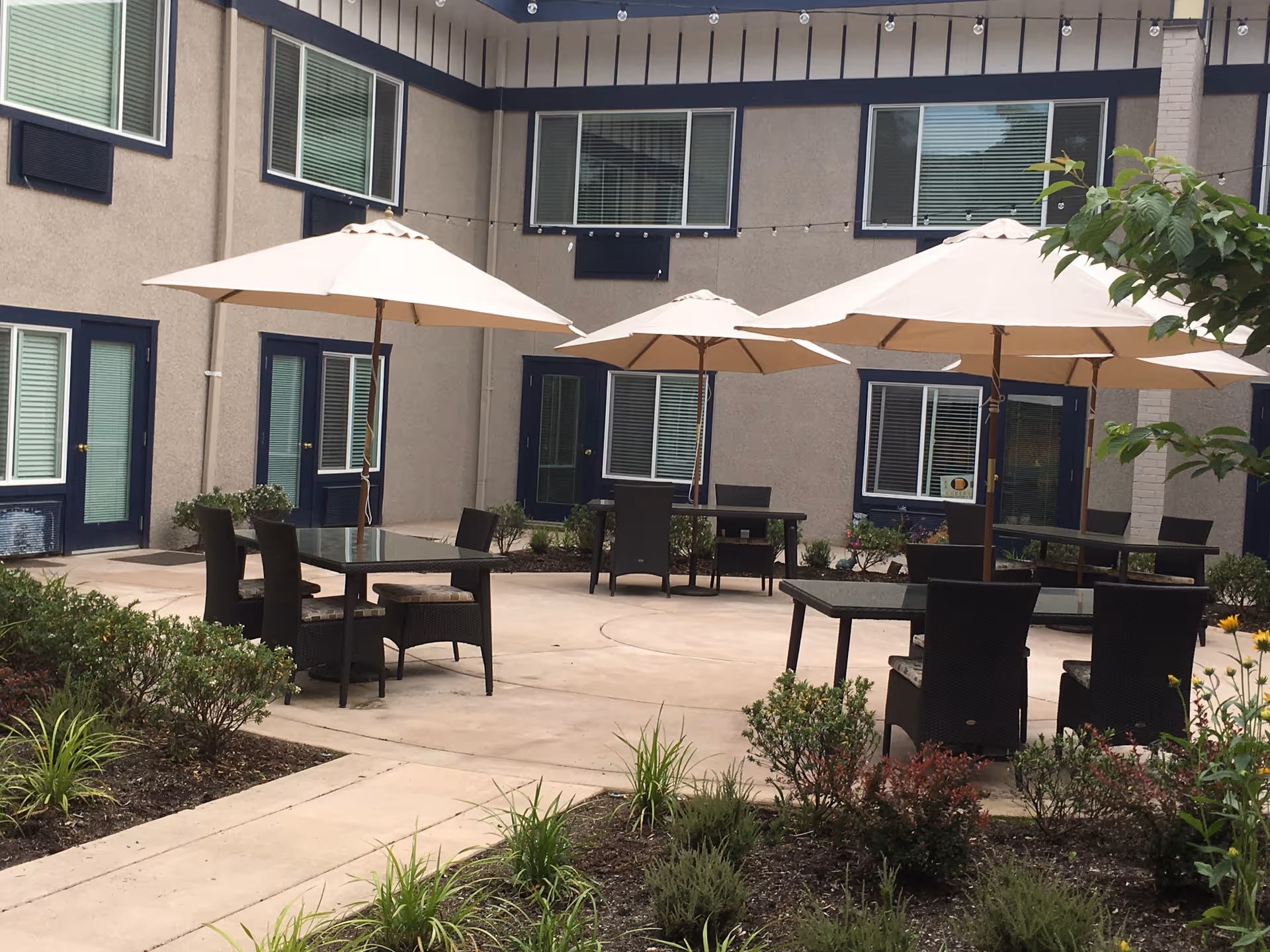 Outdoor courtyard area with several tables and chairs under large beige umbrellas. The courtyard is surrounded by a two-story building with windows and doors, and there are plants and shrubs around the paved seating area.